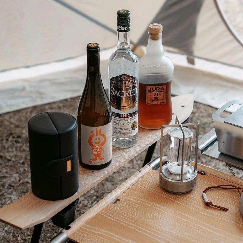 Silver cordless LED table lamp on wooden camping table with wine bottles and outdoor picnic scene, warm light