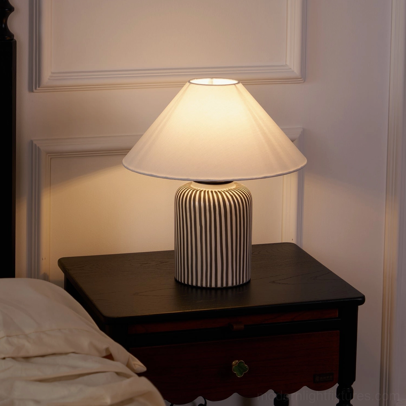 Warmly lit hand-painted striped ceramic table lamp on dark wood nightstand with wainscoting wall background
