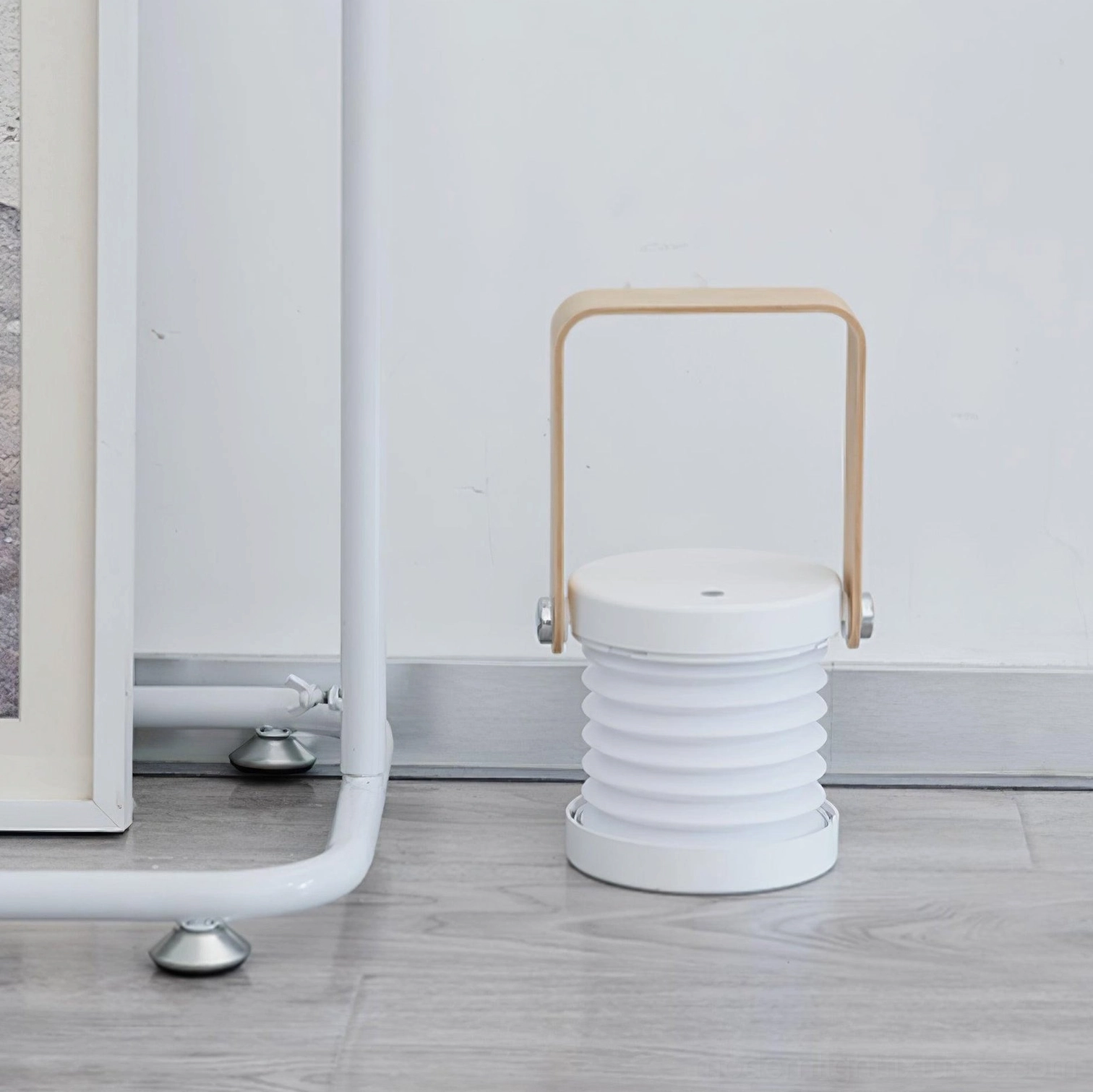 Collapsible Terna Portable Table Lamp - Rear side view showing white accordion lamp body and light wood carrying handle in indoor wooden corner
