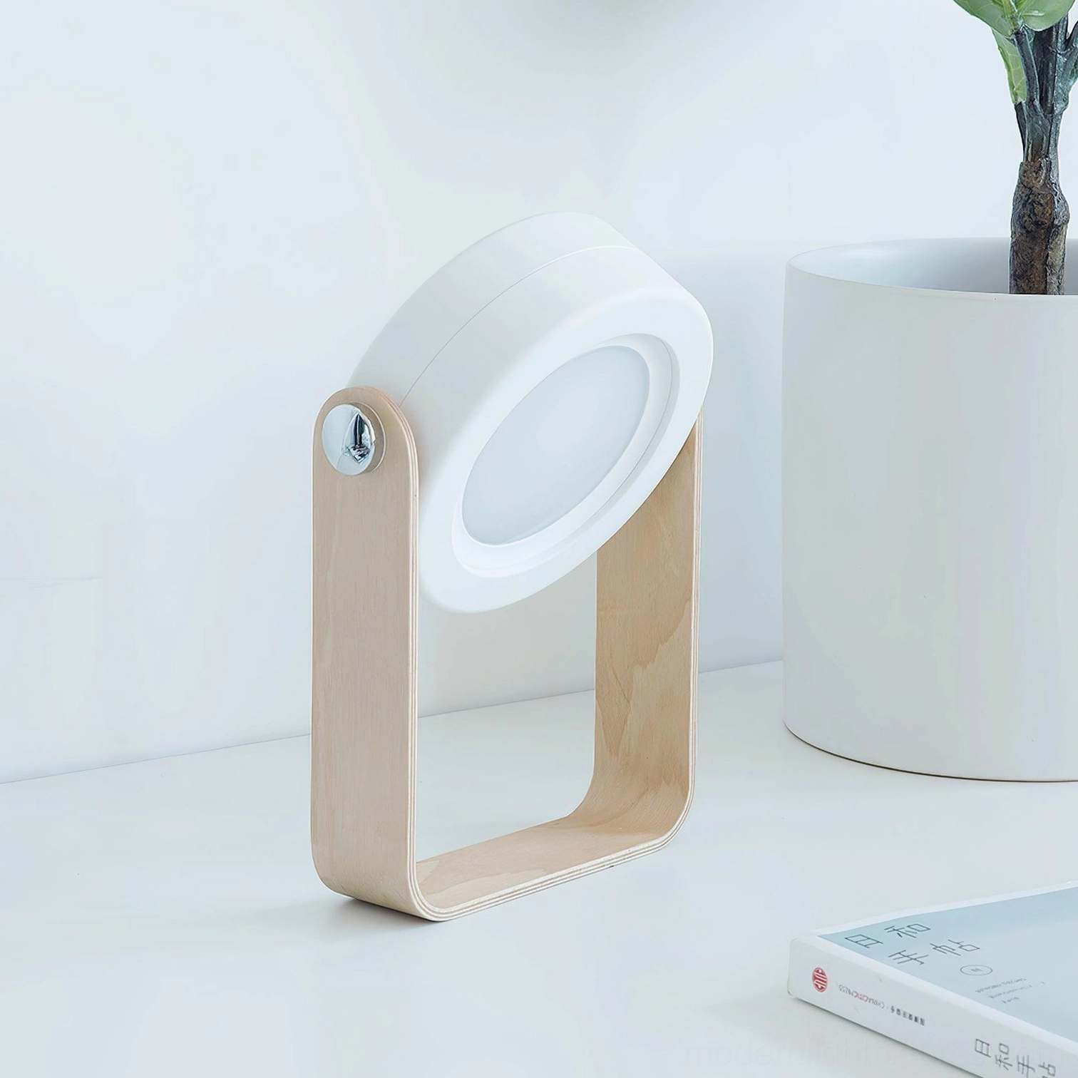 Terna Portable Table Lamp - Side three-quarter view on home desk with light wood frame, rotatable white lamp head, surrounded by book and potted plant