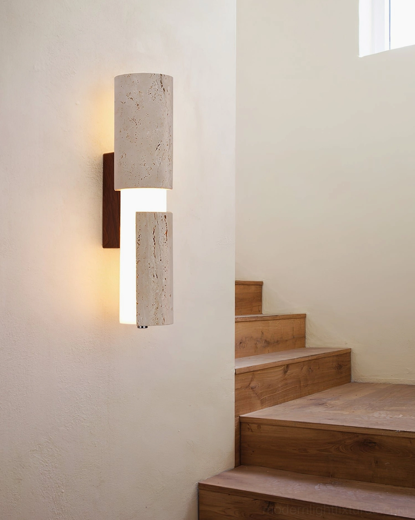 Tolira wall sconce installed in residential stairwell, side perspective showing warm ambient lighting and natural texture of travertine stone