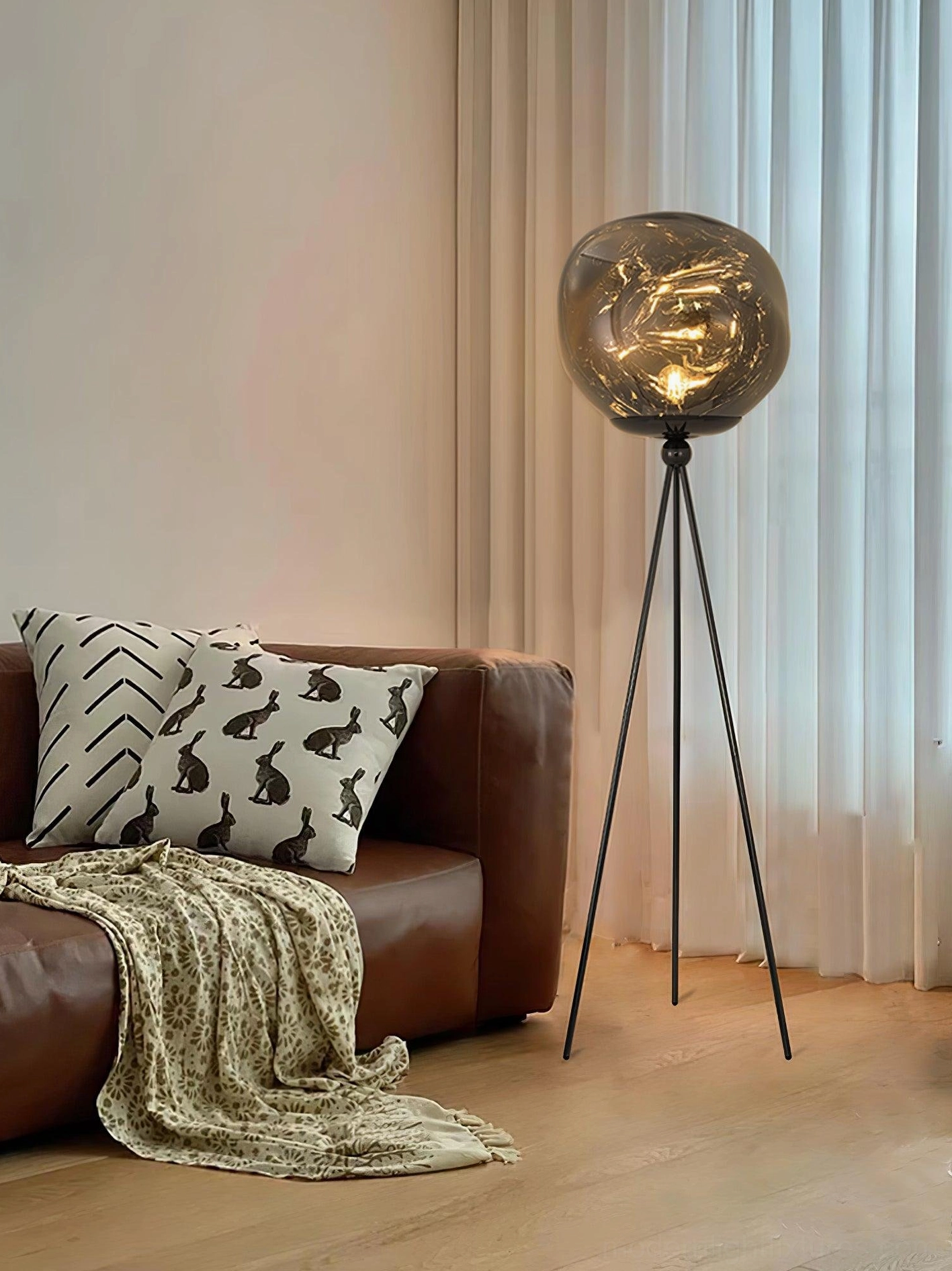 Black 3-legged tripod floor lamp with smoked glass shade in living room with brown leather sofa and sheer curtains