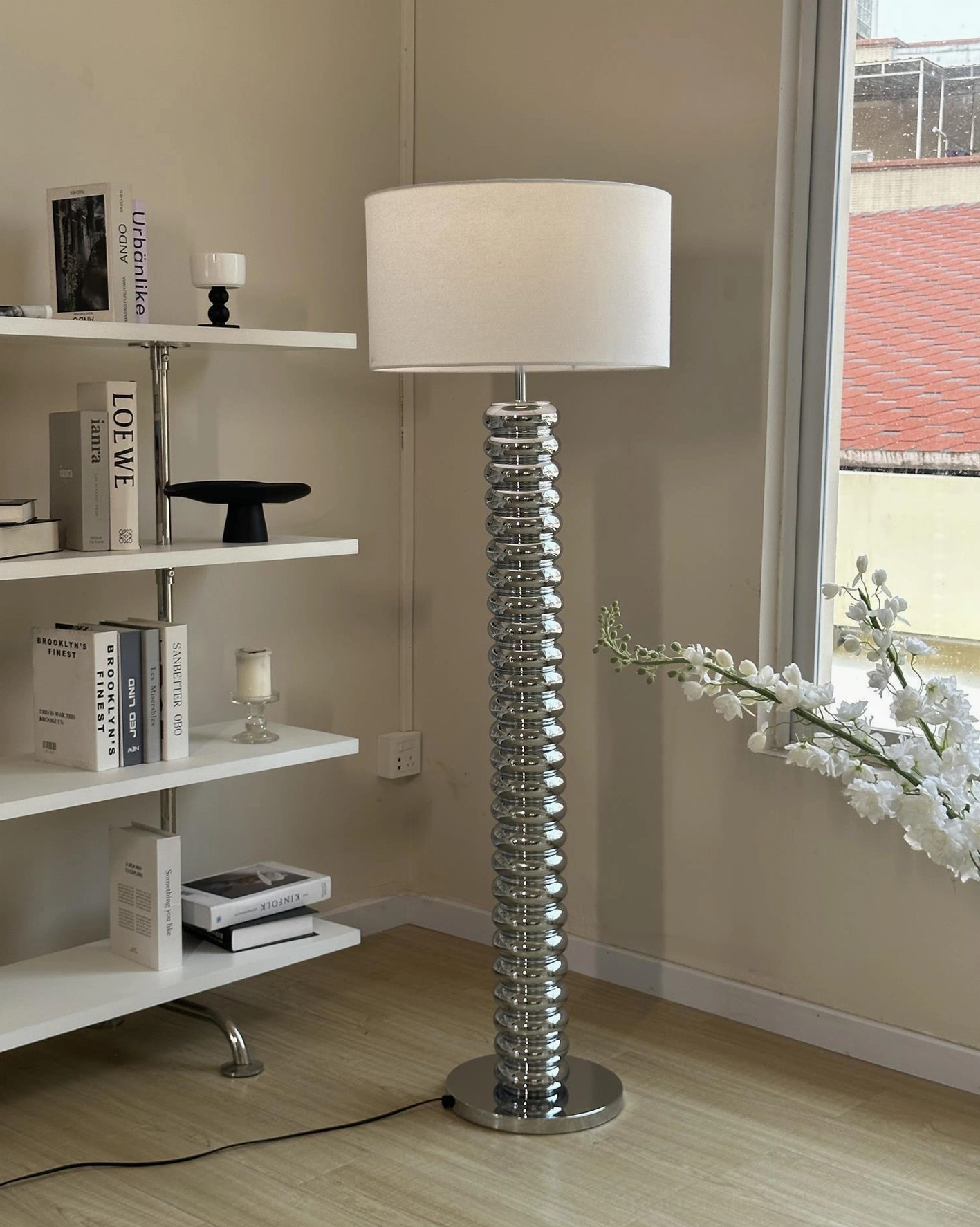 Polished chrome ribbed metal modern floor lamp with white fabric shade beside bookshelf and floral home decor