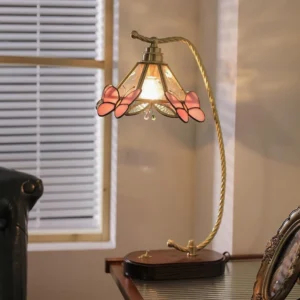 Lit Valerie Butterfly decorative table lamp on desk with louvered window, showing warm brass arm and illuminated fabric shade with pink butterfly trim