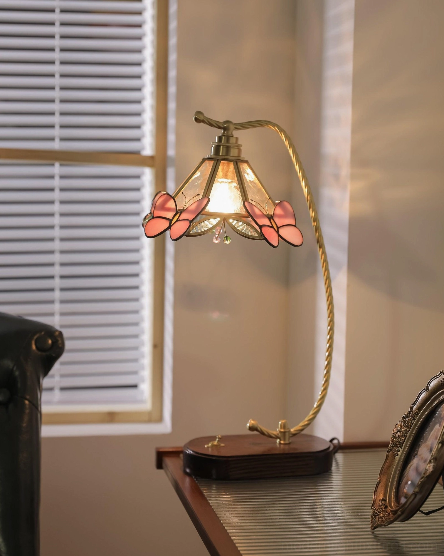 Lit Valerie Butterfly vintage table lamp on desk with louvered window, featuring warm red butterfly shade accents and glowing brass curved structure