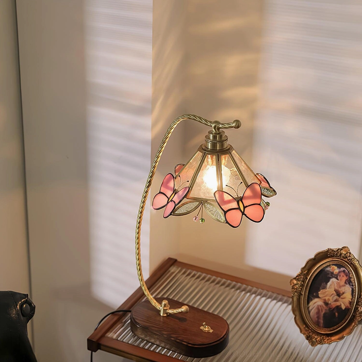 Valerie Butterfly lamp three-quarter angle on desk with ribbed glass top, showing lit pink butterfly shade accents and brass curved arm