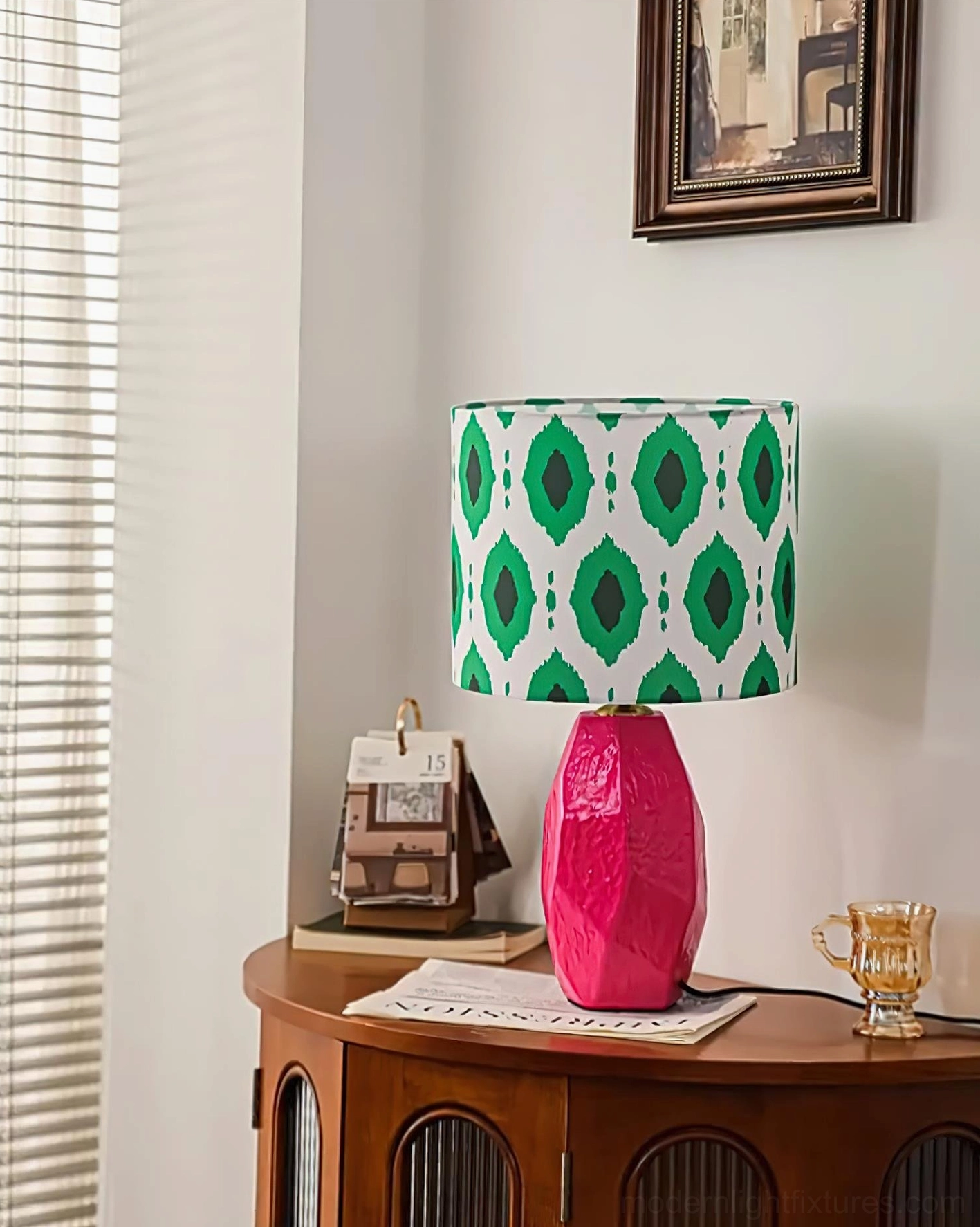 Vico geometric ikat table lamp with textured rose red ceramic base and green patterned fabric shade, three-quarter view with window scene