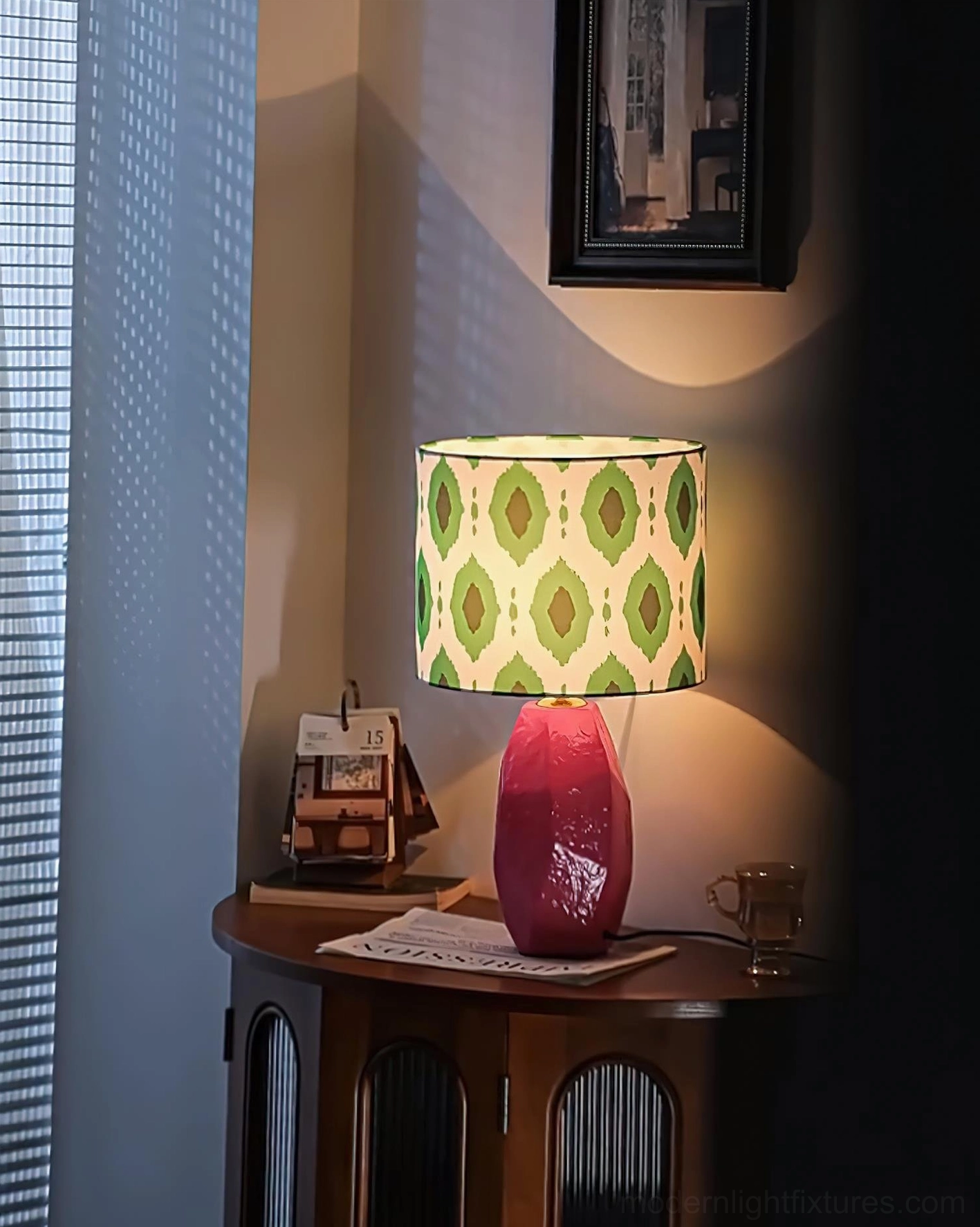 Vico modern geometric table lamp illuminated with warm light, low frontal view showing textured pink base against vintage interior with framed art