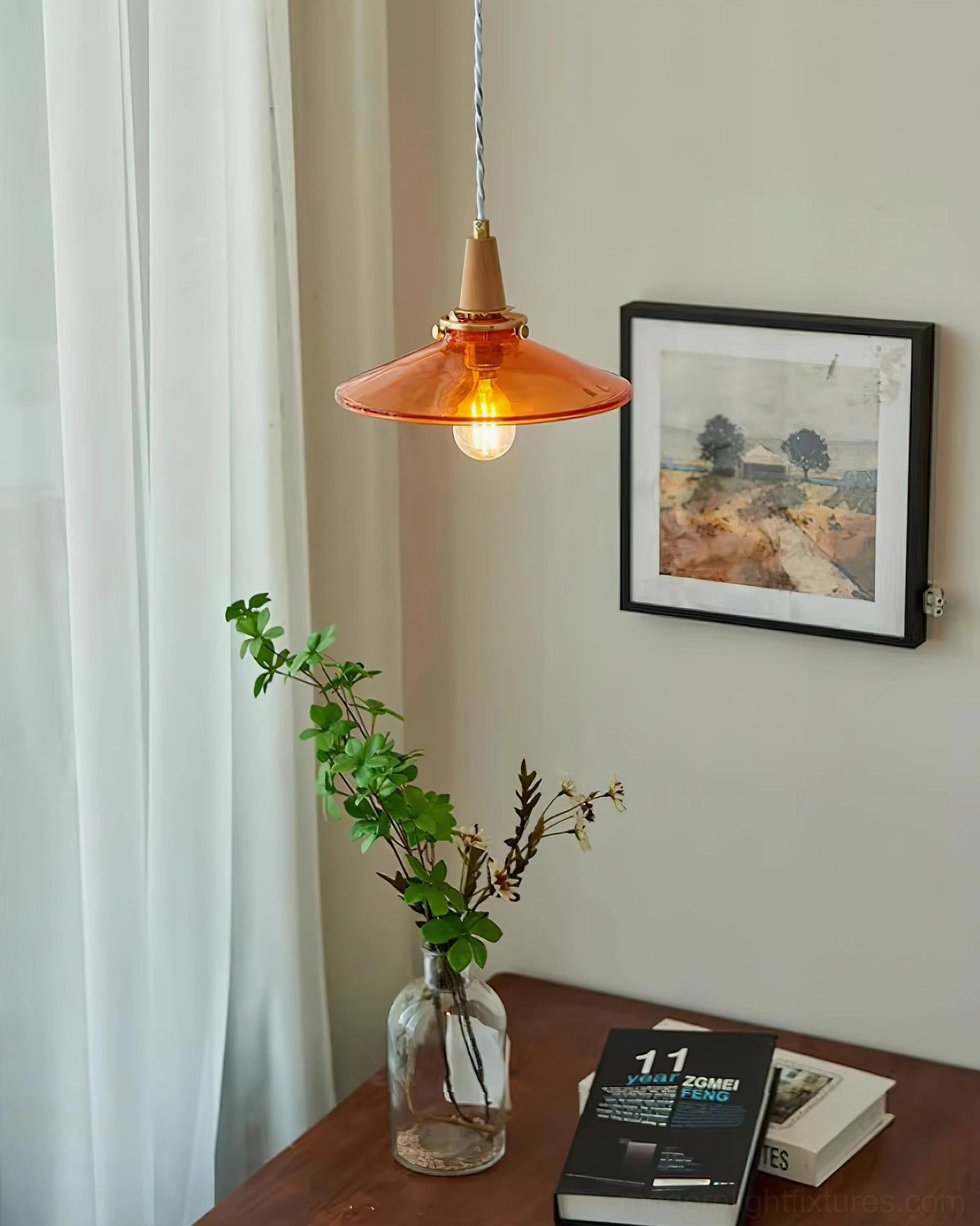 Vintage amber pendant light mounted above wooden table with stacked books, potted plant in framed corner