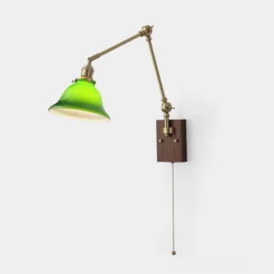 Vintage brass swing arm wall sconce with green glass dome shade on plain white wall, retro banker lamp design with dark wood backplate and pull chain switch