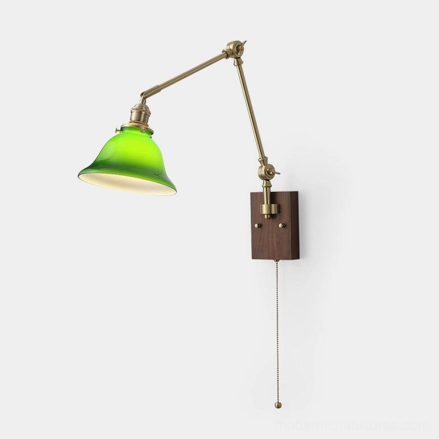 Vintage brass swing arm wall sconce with green glass dome shade on plain white wall, retro banker lamp design with dark wood backplate and pull chain switch