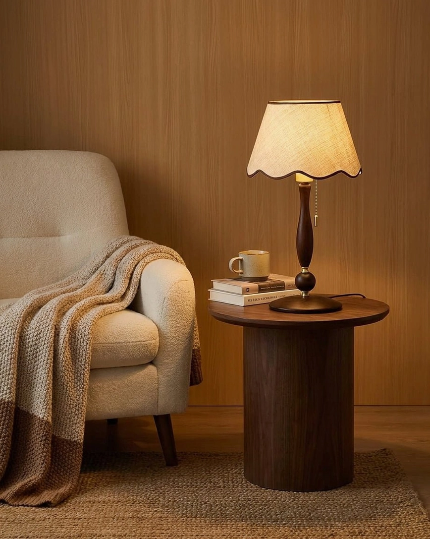 Vintage-inspired scalloped table lamp with warm glowing light next to upholstered armchair with knit throw blanket in cozy living room corner