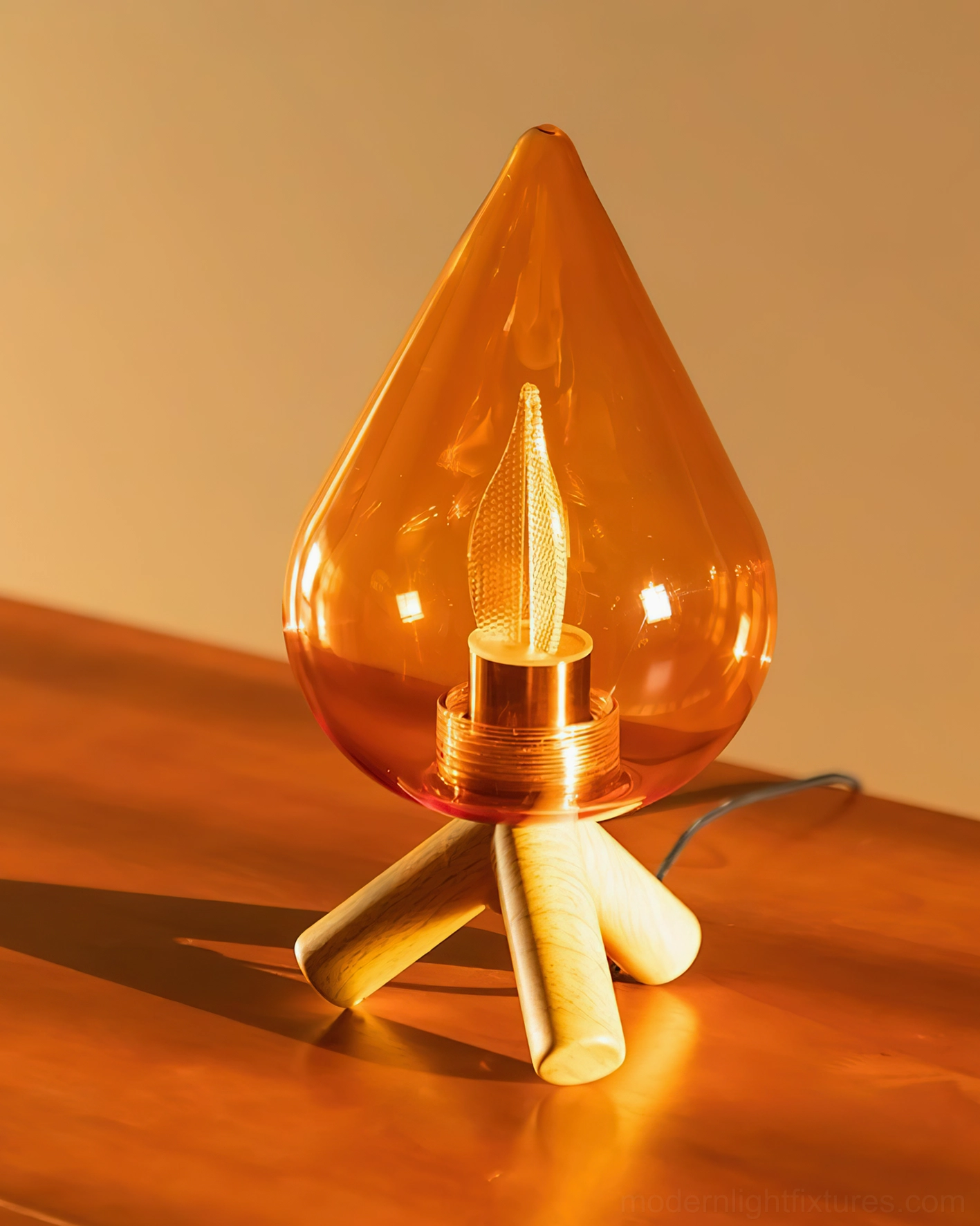 Teardrop amber glass lamp with illuminated warm bulb on solid wood tabletop, three-quarter view showing tripod base wood grain detail