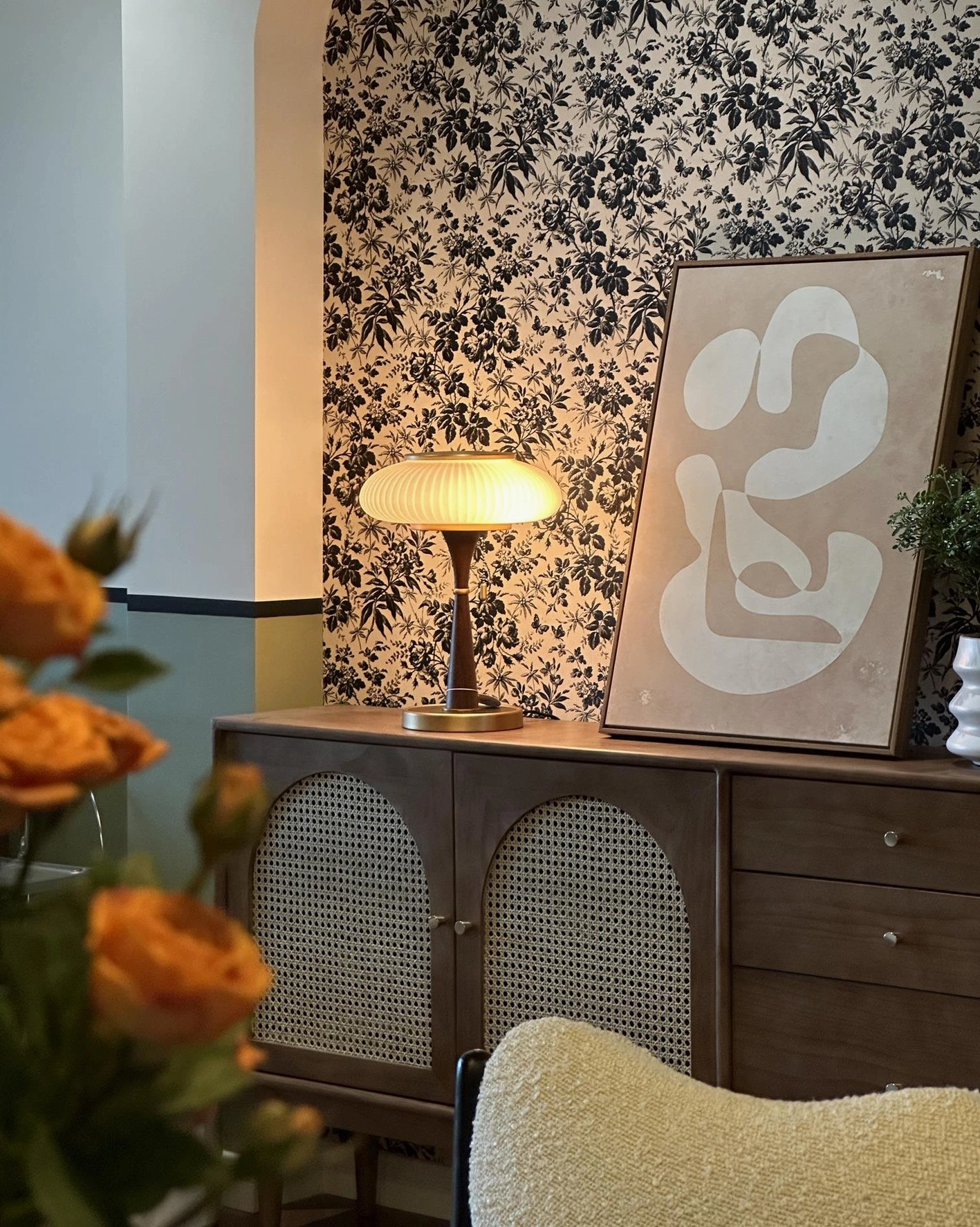 Welma mid-century modern table lamp with brass accents wood grain pole warm amber ribbed shade on decorative side cabinet with floral wallpaper