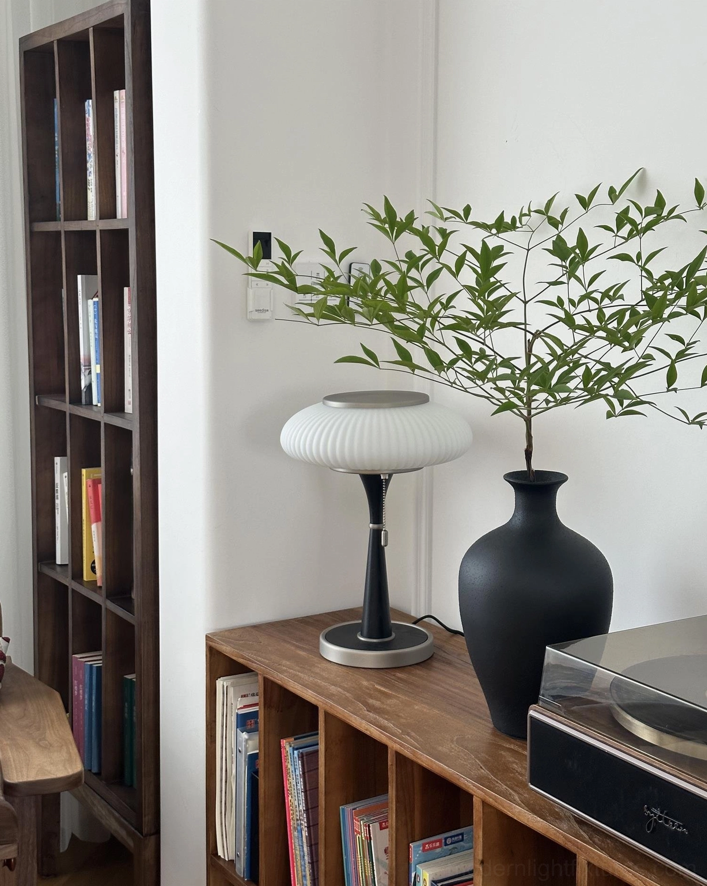 Mid-century modern pleated table lamp with black metal pole silver base and white ribbed shade on bookshelf side table with green plant