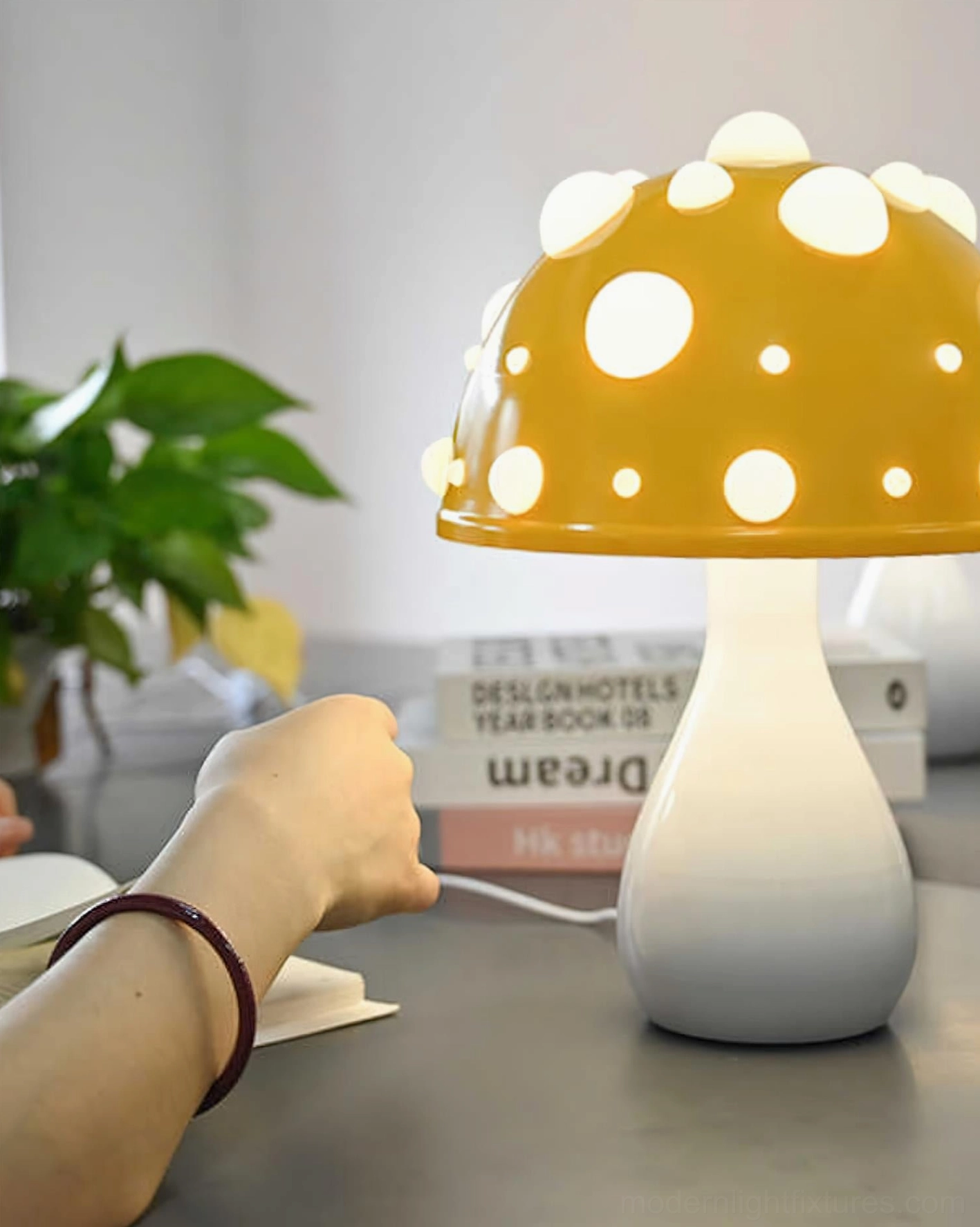 Yellow spotted mushroom table lamp with warm glowing cap on cozy home desk with green plant, three-quarter view